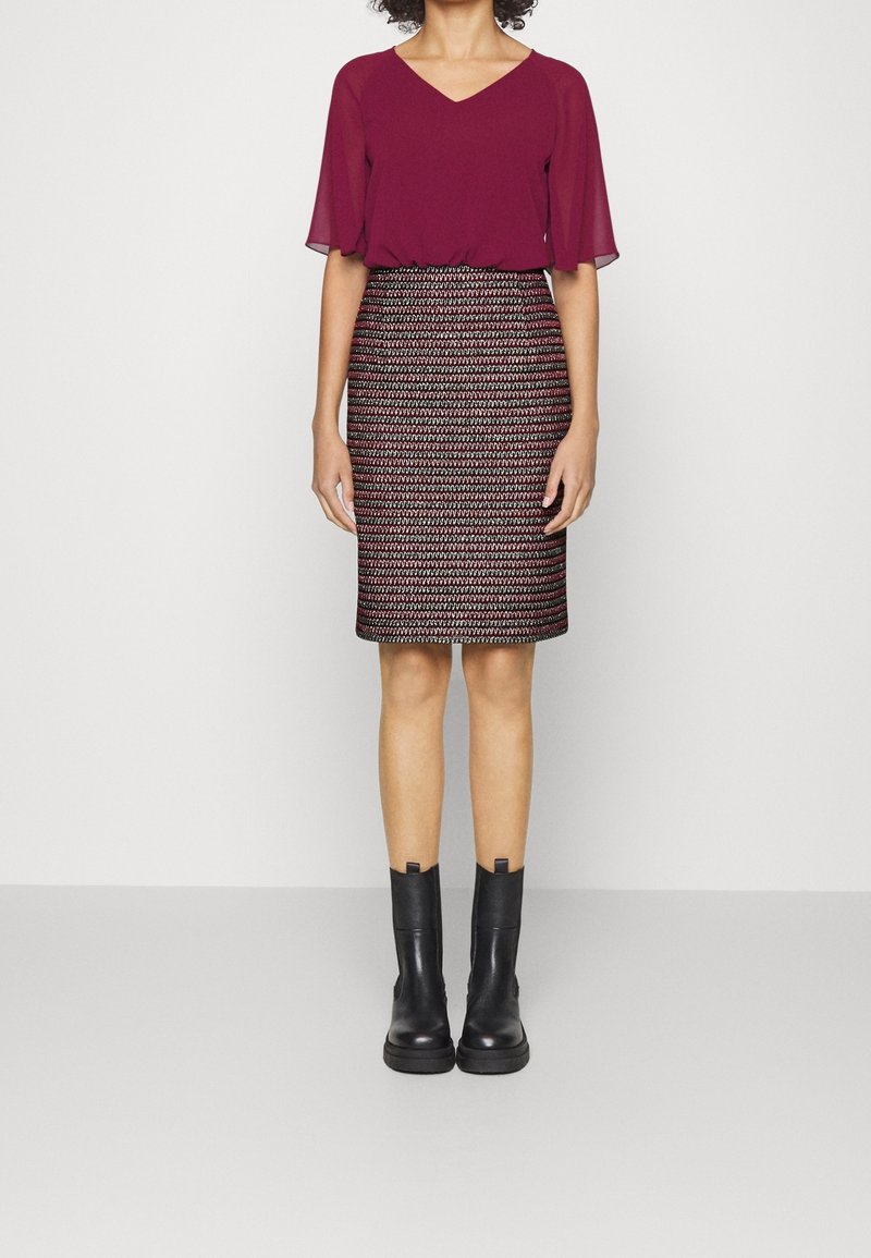 Femme portant un chemisier bordeaux à col en V, une jupe crayon à motifs mi-cuisses, et des bottines noires, debout devant un fond clair uni.
