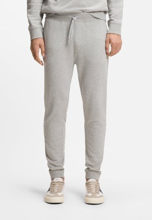 Homme portant un pantalon de jogging gris clair et des baskets beige debout sur un fond blanc.
