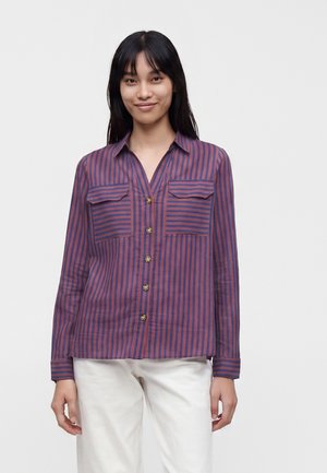Femme portant une chemise boutonnée à rayures verticales violettes et rouges avec deux poches poitrine, associée à un pantalon blanc, debout devant un fond uni.