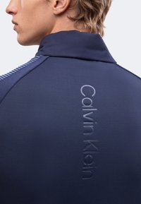 Personne blonde portant une veste bleu marine avec un logo Calvin Klein en relief, vertical, au dos près du col.