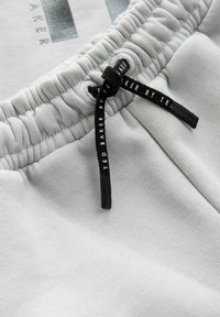 Pantalones cortos de algodón gris claro con una cintura elástica y cordón negro. Presentan el nombre "Ted Baker" impreso en el cordón.