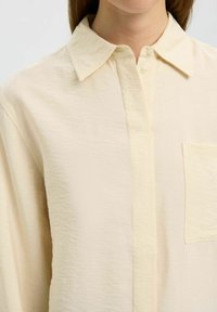 Camisa de botón de color beige claro con un acabado texturizado, un cuello clásico y un bolsillo de parche en el lado izquierdo. Tejido suave y corte oversize.