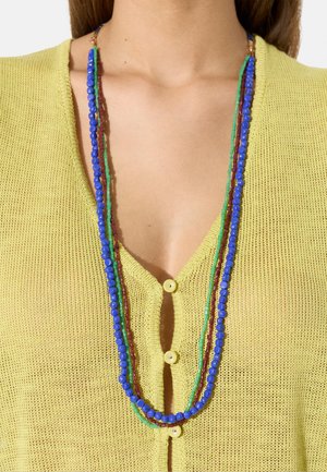 Osoba ubrana w żółty, zapinany na guziki, dziergany top oraz długi naszyjnik z wieloma rzędami niebieskich, zielonych i brązowych koralików.