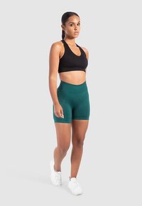 Zwarte sportbeha en groene shorts met hoge taille, gemaakt van glad, rekbaar materiaal. Model poseert tegen een lichtgrijze achtergrond.