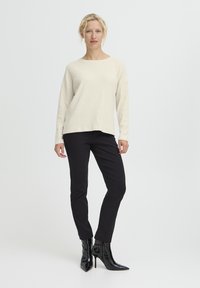 Maglione lungo a maniche lunghe color beige chiaro a coste abbinato a pantaloni neri a taglio slim e stivaletti neri lucidi con tacco alto. Sfondo semplice.