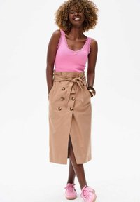 Jupe beige mi-longue avec des boutons croisés, des poches latérales et une taille à nouer. Associée à un débardeur rose et des baskets roses.