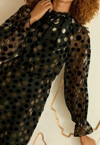 Rochie neagră transparentă cu buline mari metalice aurii și negre. Prezintă un guler cu fundă, mâneci bufante și manșete cu volane. Textură netedă.