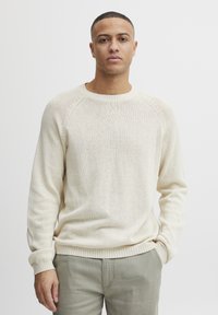 Cremefarbenes Strickpullover mit Rundhalsausschnitt, gerippten Bündchen und Saum; leichte Textur mit einem subtilen Muster. Kombiniert mit hellen Hosen.