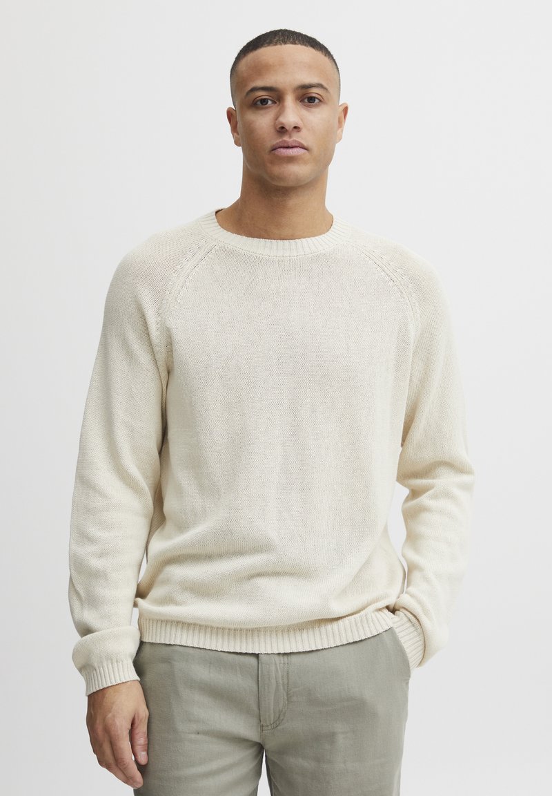 Cremefarbenes Strickpullover mit Rundhalsausschnitt, gerippten Bündchen und Saum; leichte Textur mit einem subtilen Muster. Kombiniert mit hellen Hosen.