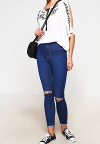 Blouse blanche avec broderie florale noire, associée à un jean skinny bleu avec des genoux déchirés, et des baskets noires avec des lacets blancs.