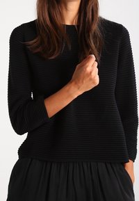 Haut noir texturé avec des côtes horizontales, manches trois-quarts et coupe ample. Détails comprenant un col rond et un ourlet superposé.