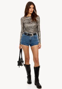 Haut à manches longues argenté métallisé, shorts en denim, ceinture noire avec des clous, bottes noires jusqu'aux genoux et petit sac à main noir.