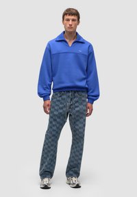 Felpa blu con colletto corto, abbinata a pantaloni in denim con stampa all-over del logo "WB". Completa con scarpe da ginnastica.