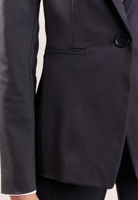 Blazer nero sartoriale con chiusura a un bottone, rever strutturato e una elegante texture in satin, con dettaglio della tasca laterale.
