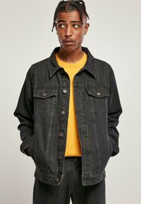 Ung mann med flettet hår iført en svart denimjakke over en gul t-skjorte, med hendene i lommene, og ser til siden.