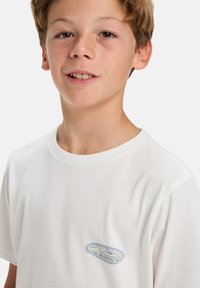 Hvit bomulls t-skjorte med rund hals og korte ermer. Har en liten blå brodert logo på venstre brystområde.