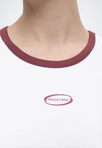 Κοντινό πλάνο ενός λευκού πουκαμίσου με μπουρνουζολεπτό μαρόν γιακά και ένα μικρό μαρόν λογότυπο με την επιγραφή "MODSTRÖM" στο στήθος.