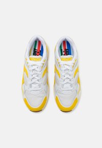 Weiße Sportschuhe mit gelben Akzenten, bestehend aus einem Obermaterial aus Mesh und Wildleder, Schnürverschluss und einer strukturierten Gummisohle. Buntes Fußbett sichtbar.