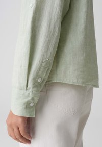 Camicia di lino verde chiaro con finitura testurizzata, dotata di polsini con bottoni e orlo curvo, abbinata a pantaloni chiari in stile denim.