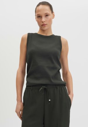 Donna con top scuro senza maniche e pantaloni con coulisse abbinati, in piedi con una mano in tasca su sfondo uniforme.