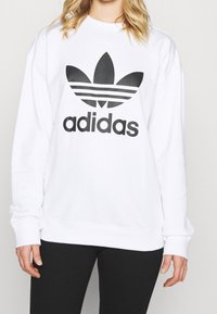 Vit sweatshirt med en stor svart Adidas-logotyp med trebladig design och tre horisontella ränder, tillverkad av mjukt bomullsmaterial.