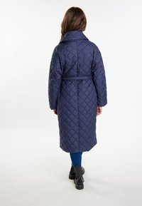 Marineblå quiltet lang jakke med bælte i taljen, stor krave og nedsat kant. Modellen har blå leggings og sorte ankelstøvler på.