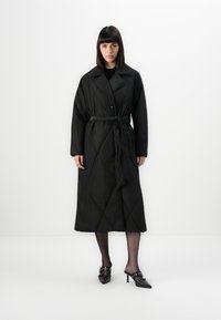 Woman with straight dark hair wearing a long black quilted coat with a belt, black tights, and black pointed-toe heeled mules with buckles.