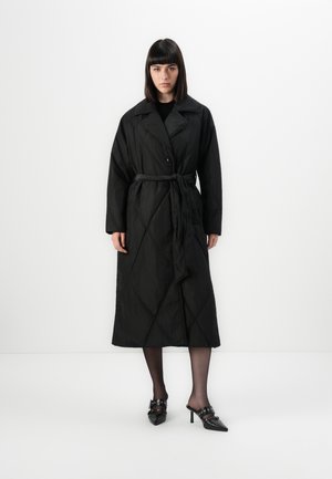 Woman with straight dark hair wearing a long black quilted coat with a belt, black tights, and black pointed-toe heeled mules with buckles.