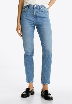 Person trägt hellblaue, hochtaillierte Jeans mit geradem Bein, schwarze Loafers und ein weißes, in die Hose gestecktes Oberteil, steht vor einem schlichten Hintergrund.