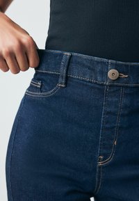 Tmavě modré džíny z denimu se přiléhavým pasem, vybavené zapínáním na knoflík, kontrastním prošitím a malým detailem přední kapsy.