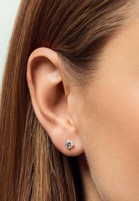 Klart studörhänge med en rund, glittrande diamantliknande centersten, placerad i en enkel metallbas, bärs på en modells öra.