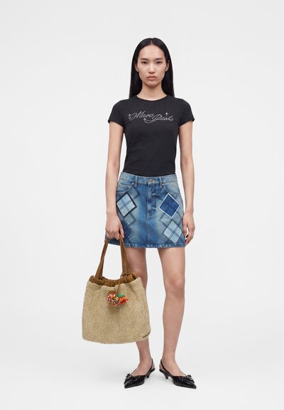 Νεαρή γυναίκα που φοράει μαύρο εφαρμοστό t-shirt με σενάριο λογότυπο, μπλε τζιν μίνι φούστα με διαμαντένια μπαλώματα, κρατώντας πλεκτό τσάντα tote με κρέμασμα πομ πομ.
