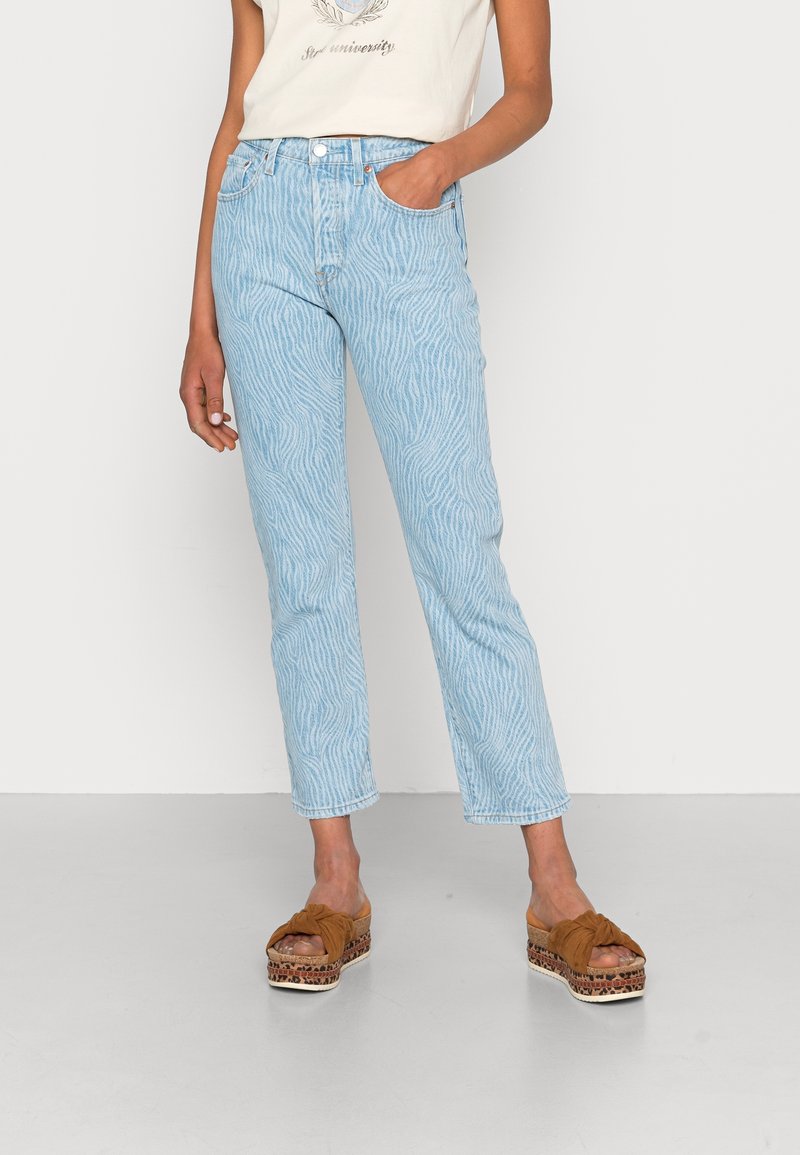 Jean taille haute bleu clair avec un motif ondulé subtil, porté avec des sandales compensées couleur tan à brides nouées, sur une personne debout.