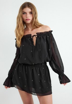 Młoda kobieta z długimi blond włosami, ubrana w czarną sukienkę odkrywającą ramiona, z przezroczystymi rękawami i małymi okrągłymi ozdobami.