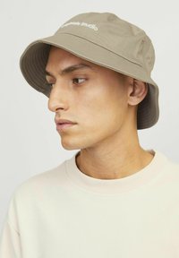Beige bucket hat with a wide brim, made of cotton, featuring stitched detailing and a white logo printed on the front.
