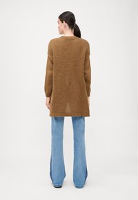 Suéter marrón de punto con un corte largo y relajado, puños acanalados y una abertura en la parte trasera central. Combinado con jeans acampanados de color azul claro.
