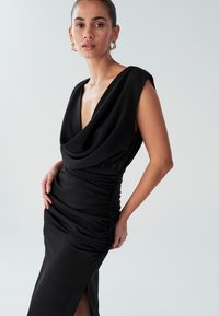 Abito nero con scollatura a V profonda, corpetto drappeggiato e dettagli arricciati. Presenta un design senza maniche e una spaccatura laterale per facilità di movimento.