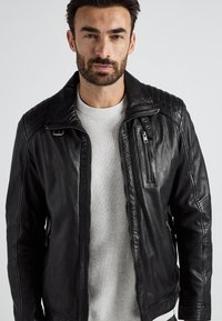 Svart läderjacka med quiltat axeldesign, dragkedja framtill och en liten bröstficka. Bärs över en ljusgrå tröja.