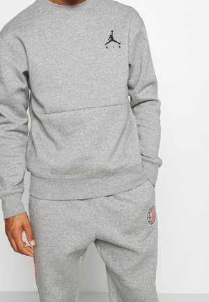 Mężczyzna ubrany w szarą bluzę z kapturem i pasujące spodnie dresowe z czarno-czerwonymi logo Jumpman na piersi i górnej części uda.