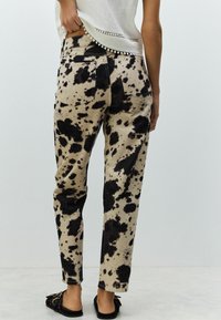 Jean beige délavé avec imprimé animal noir, coupe ajustée et cinq poches, associé à des chaussures plates noires. Détails visibles à la taille et à l'ourlet.