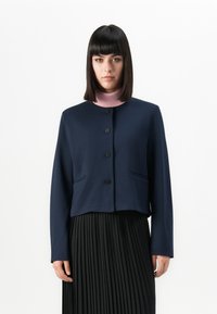 Femme avec des cheveux noirs lisses portant une veste bleu marine à boutons au-dessus d'un col roulé rose et d'une jupe plissée noire sur un fond blanc uni.