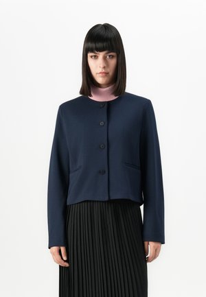 Vrouw met steil zwart haar, gekleed in een marineblauwe button-up jas over een roze coltrui en een zwarte plissérok tegen een effen witte achtergrond.