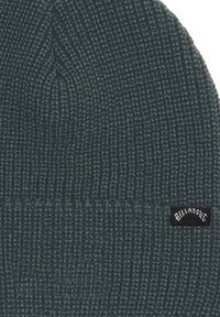 Berretto lavorato a maglia di colore verde scuro, con una texture a coste e un risvolto pieghevole. Presenta un piccolo etichetta nera con testo bianco sul lato.