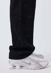 Pantaloni neri con un sottile motivo a logo tono su tono, abbinati a sneaker Nike bianche con un accento argento e un design distintivo di ammortizzazione a molla.
