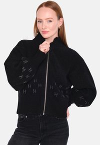 Svart zip-up tröja med hög krage, oversized ärmar och ribbade muddarna. Har ett subtilt logomönster på den nedre framsidan. Mjuk textur.