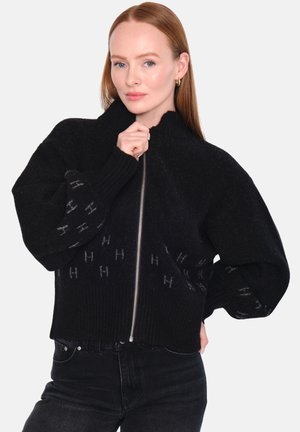 Svart zip-up tröja med hög krage, oversized ärmar och ribbade muddarna. Har ett subtilt logomönster på den nedre framsidan. Mjuk textur.