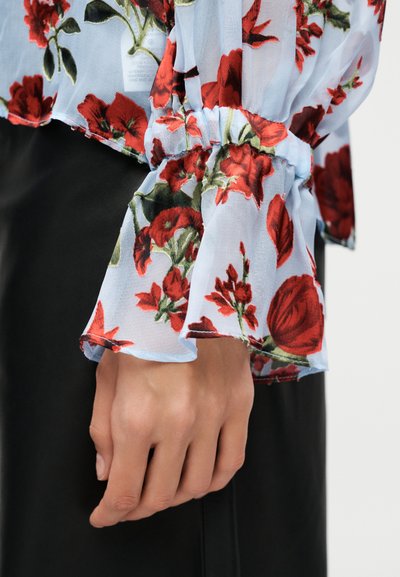Χέρι που ακουμπάει στο πλευρό, φορά διαφανή ανοιχτό μπλε μπλούζα με κόκκινο ανθισμένο μοτίβο και συγκεντρωμένο βολάν στο μανίκι πάνω από μαύρο παντελόνι.