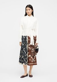 Γυναίκα που στέκεται φορόντας λευκό πουκάμισο με ζώνη και μεσαίο φούστα με σκίσιμο με μαύρα και καφέ floral σχέδια, συνδυασμένο με σκούρα καφέ επίπεδα παπούτσια.