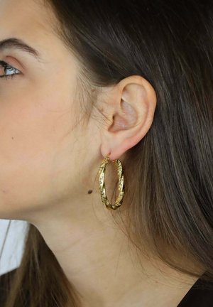 Złote kolczyki w kształcie koła z teksturowanym, skręconym wzorem. Kolczyki są duże i okrągłe, co podkreśla ich widoczny profil.