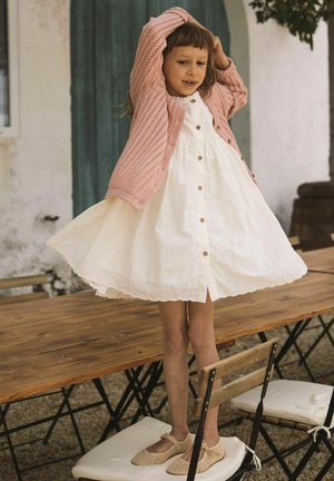 Ein junges Mädchen steht auf einem Holzstuhl, trägt ein weißes Kleid und eine rosa Strickjacke und hält ihre Hände im Freien über ihren Kopf.